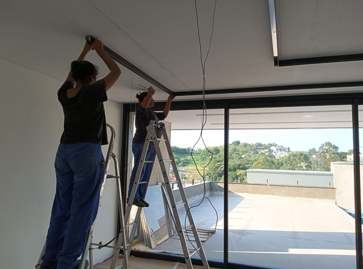 Dois eletricistas da J&C sobre escadas, fixando o perfil de uma luminária linear no teto de um ambiente com vidraça e vista para fora.