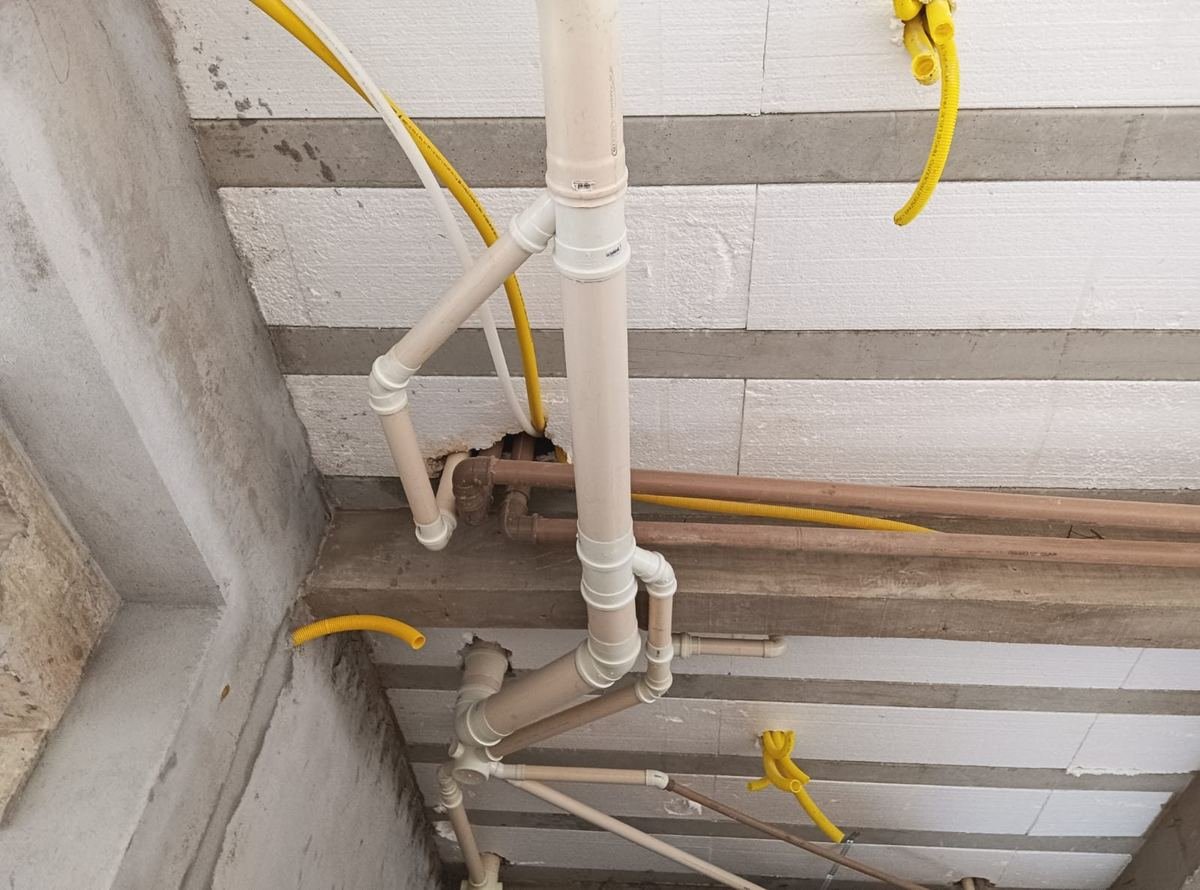 Tubulação de esgoto em PVC descendo da laje, com ramais de água ao lado e conexões montadas, antes do fechamento do forro, executada pela J&C Elétrica.